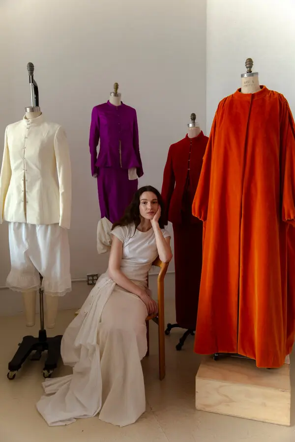 Colleen Allen sits thoughtfully on a chair, wearing a stylish white dress. Behind her are mannequins showcasing vibrant outfits, including a white top with matching pants, a purple set, a red jacket, and a large, flowing orange garment. The bright, minimalistic setting enhances the modern fashion display.